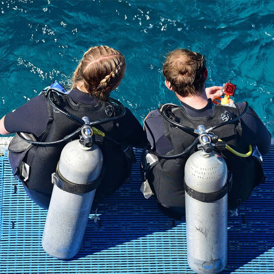 Two scuba divers sitting on a dock with their equipment.